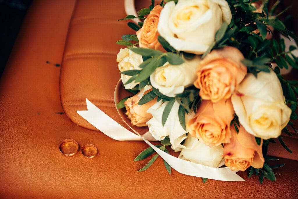 Hochzeitsstrauß aus weißen und orangen Rosen mit goldenen Trauringen auf braunem Hintergrund.